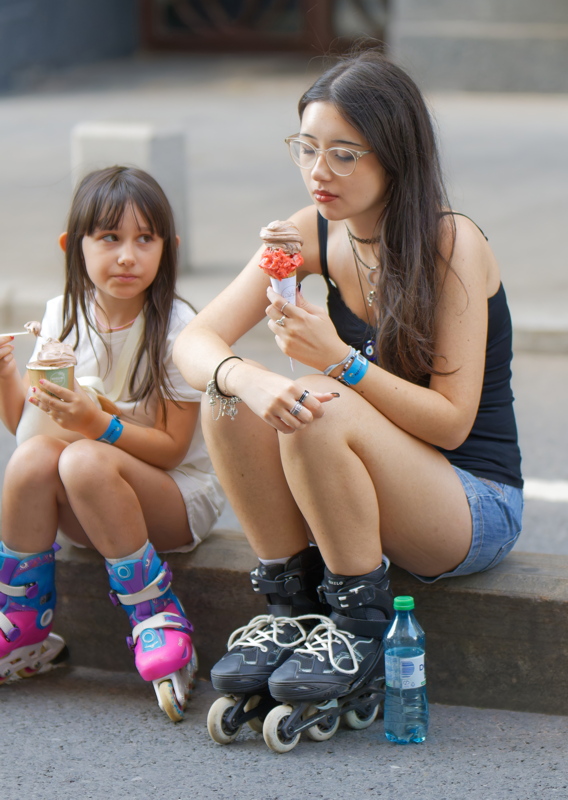23 - Two individuals eating ice cream while sitting on a curb, both wearing inline skates